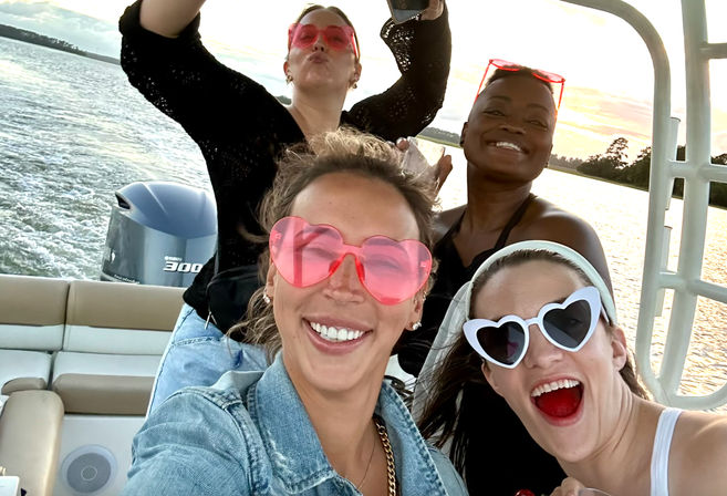 Four friends on a motorboat at sunset wearing heart-shaped sunglasses, smiling and posing for a playful selfie with water and shoreline behind them.