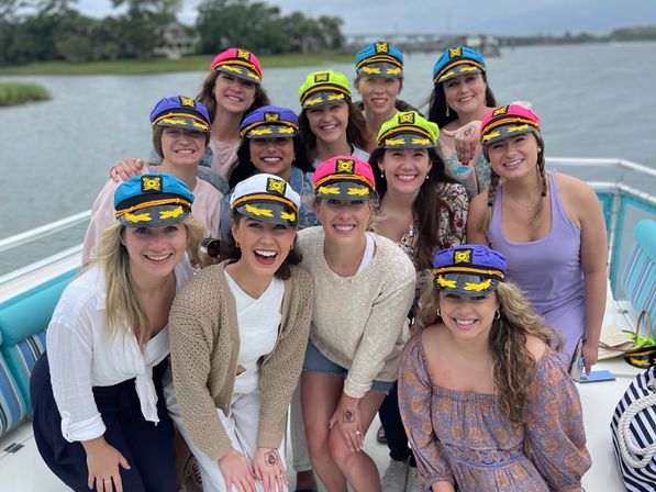 Smiling group of women in colorful captain caps posing on a boat near a coastal inlet, enjoying a daytime waterfront celebration.