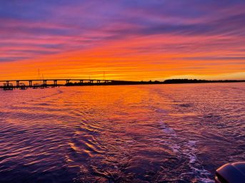 Vibrant purple-orange sunset over a coastal bay, silhouetted bridge and docks on the horizon, rippled water reflecting the colors with a boat wake in the foreground.