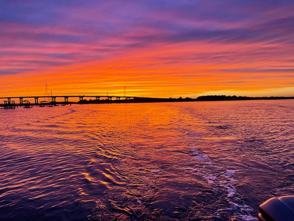 Vibrant purple-orange sunset over a coastal bay, silhouetted bridge and docks on the horizon, rippled water reflecting the colors with a boat wake in the foreground.