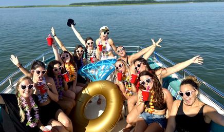 Group of friends on a sunny lake boat party, wearing heart-shaped sunglasses and leis, cheering with red cups around a gold inflatable ring and oversized diamond float
