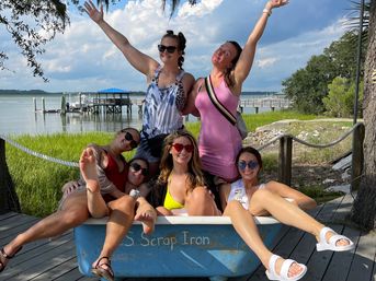 Six friends in summer outfits and sunglasses pose joyfully—some with arms raised—sitting in a blue tub on a wooden waterfront dock by marsh grasses and a boat pier under a sunny sky.