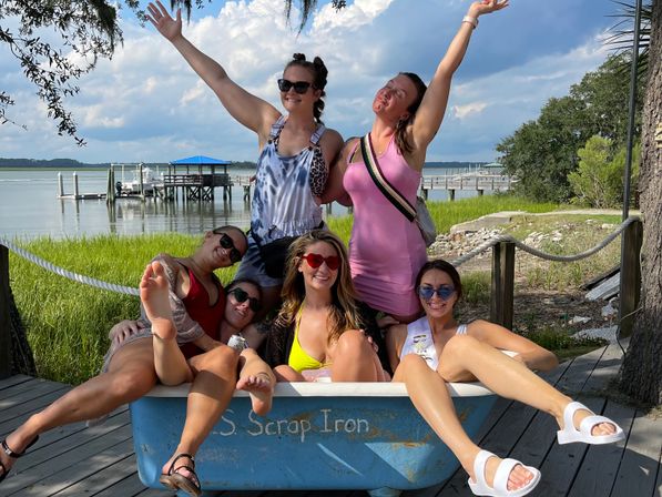 Six friends in summer outfits and sunglasses pose joyfully—some with arms raised—sitting in a blue tub on a wooden waterfront dock by marsh grasses and a boat pier under a sunny sky.