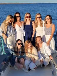 Eight friends in summer outfits smiling on a boat deck with calm blue ocean and a distant shoreline at golden-hour sunset