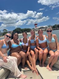 Six women in blue and white swimsuits smiling together on a boat by a sandy beach and red-and-white lighthouse under a bright, sunny sky with puffy clouds.