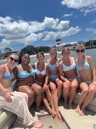 Six women in blue and white swimsuits smiling together on a boat by a sandy beach and red-and-white lighthouse under a bright, sunny sky with puffy clouds.