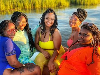 Group of five friends in bright swimsuits smiling on a small boat at golden hour with marsh grasses and calm water