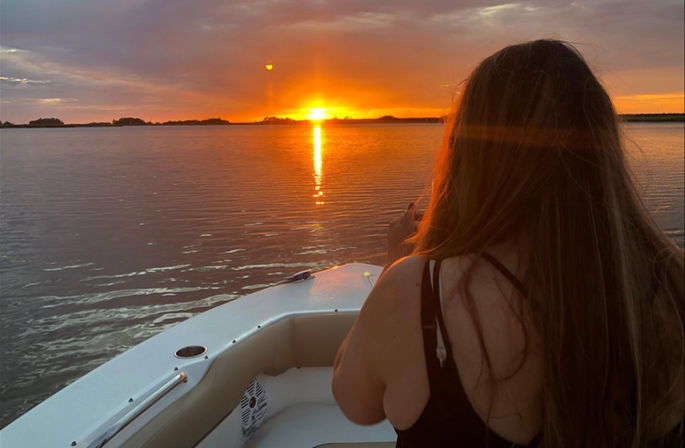 Person with long hair sitting on a boat bow watching a vibrant orange sunset reflecting across calm lake waters, golden glow on the horizon.