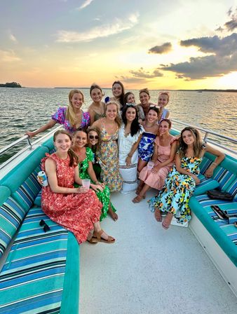 Cheerful group of women in colorful summer dresses posing on a teal-striped boat bench during a golden sunset over calm coastal waters — relaxed sunset cruise vibe.