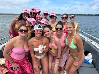 Group of smiling women in colorful bikinis and matching pink caps, holding drinks and posing together on a boat over calm coastal waters under a partly cloudy sky.