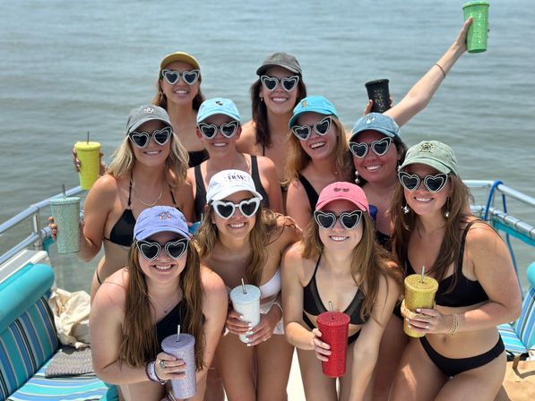 Group of women in swimsuits and baseball caps wearing heart-shaped sunglasses, holding colorful glittery tumblers and smiling on a sunny boat over calm coastal water — fun summer boat party vibe.