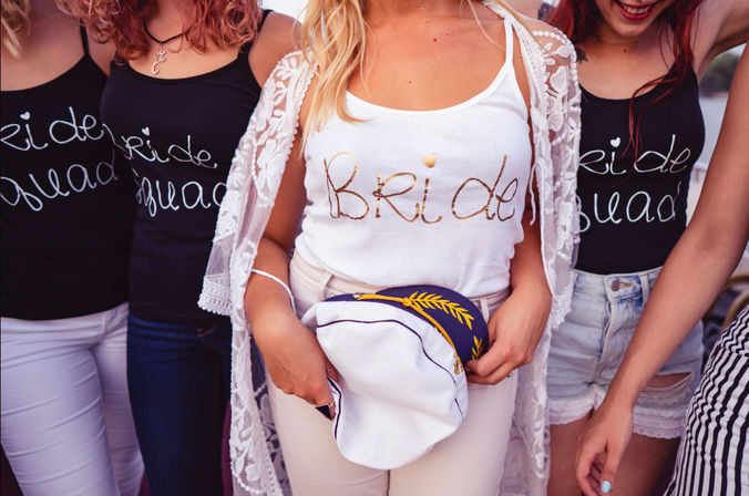 Outdoor bachelorette party: bride in a white "Bride" tank and lace cover-up holding a nautical captain's hat, flanked by bridesmaids in black "Bride Squad" tanks and casual shorts.