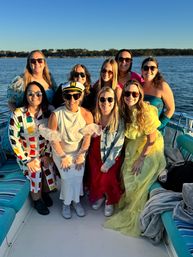 Group of women in colorful dresses and sunglasses smiling on a boat at sunset, one wearing a captain's hat, with calm water and a tree-lined shoreline in the background — sun-kissed boat party vibe.
