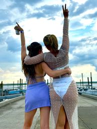 Two friends with arms around each other celebrating on a marina dock, one wearing a sheer pearl-embellished dress over white swim bottoms and the other in a blue skirt, raising hands toward a dramatic cloudy sky above boats and waterfront.