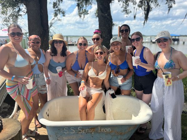 Smiling group of women in swimsuits and summer outfits holding drinks around a vintage clawfoot tub on a sunny lakeside dock with boats and trees