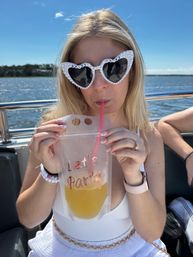 Person on a sunny boat ride wearing pearl heart-shaped sunglasses reading 'Bride to Be', sipping a yellow drink through a straw from a pouch printed 'Let's Party', with water and shoreline in the background.