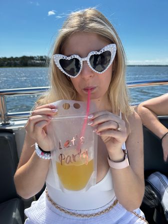 Person on a sunny boat ride wearing pearl heart-shaped sunglasses reading 'Bride to Be', sipping a yellow drink through a straw from a pouch printed 'Let's Party', with water and shoreline in the background.