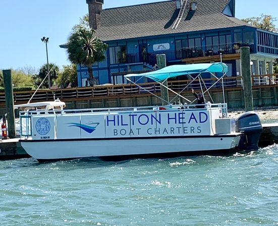Charter pontoon boat with teal canopy docked at a Hilton Head marina, palm tree and waterfront house in the background