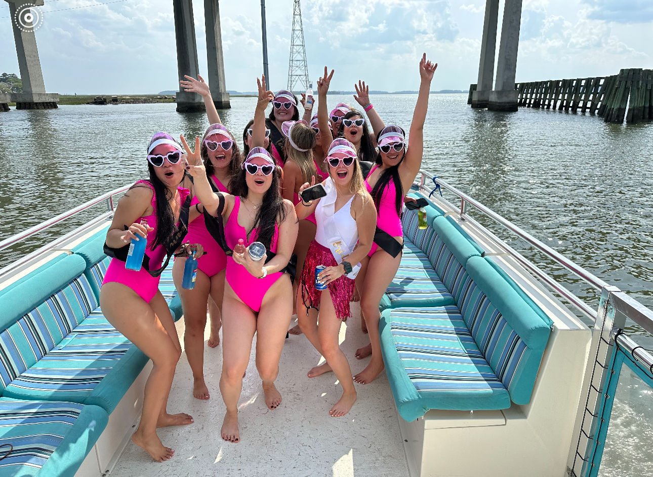 Group of women in matching hot-pink swimsuits and heart-shaped sunglasses celebrating on a party boat under a concrete bridge, holding drinks on a sunny waterfront