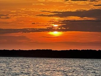 Vibrant orange and gold sunset over rippling water with the sun peeking through layered clouds above a dark silhouetted shoreline
