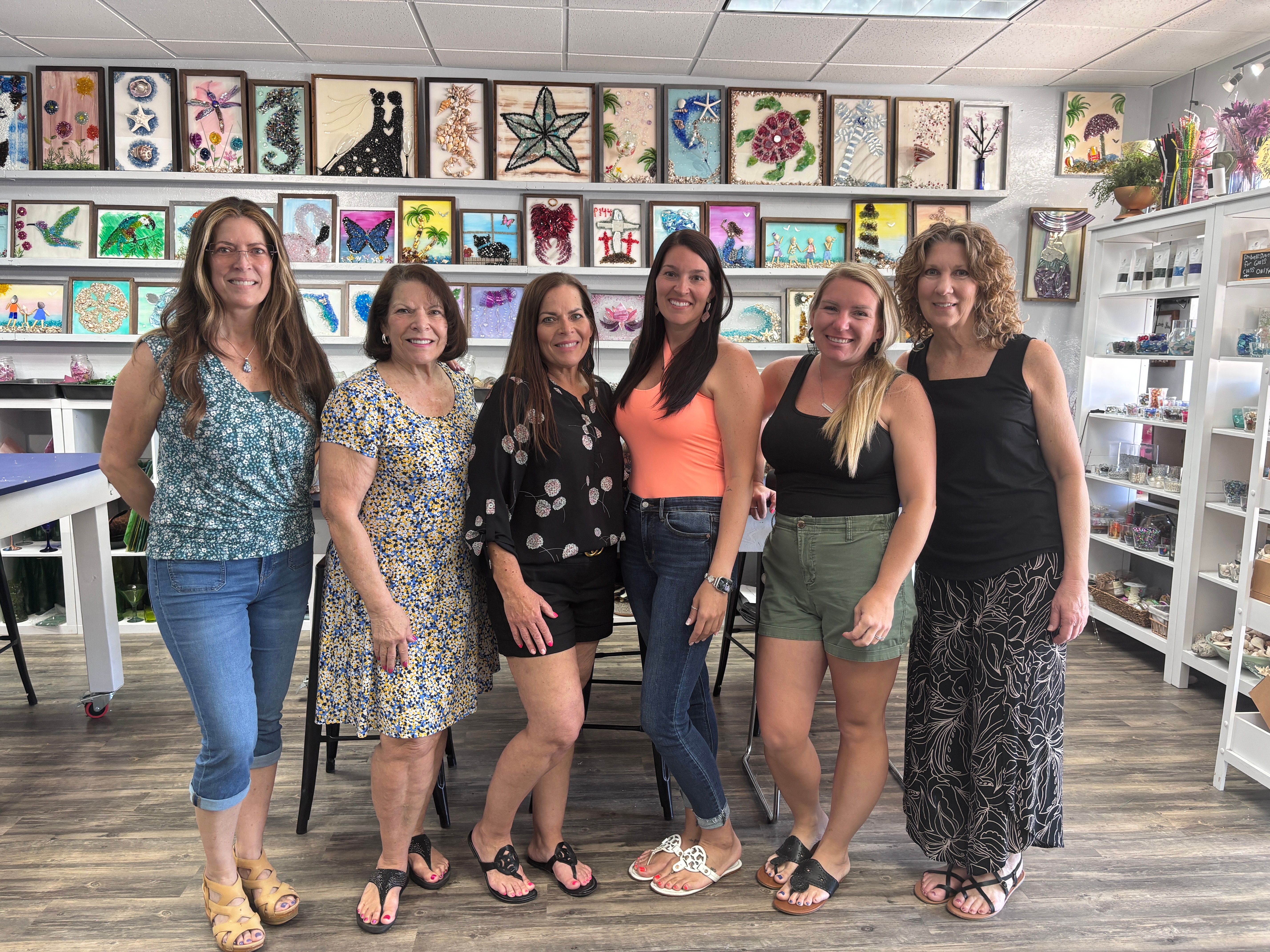 Six smiling women posing inside a bright community art studio, standing in front of shelves of colorful framed glass mosaics and mixed-media wall art, wearing casual summer outfits.