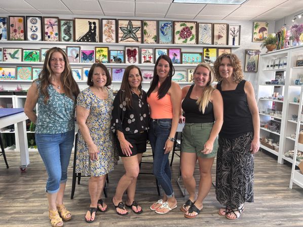 Six smiling women posing inside a bright community art studio, standing in front of shelves of colorful framed glass mosaics and mixed-media wall art, wearing casual summer outfits.
