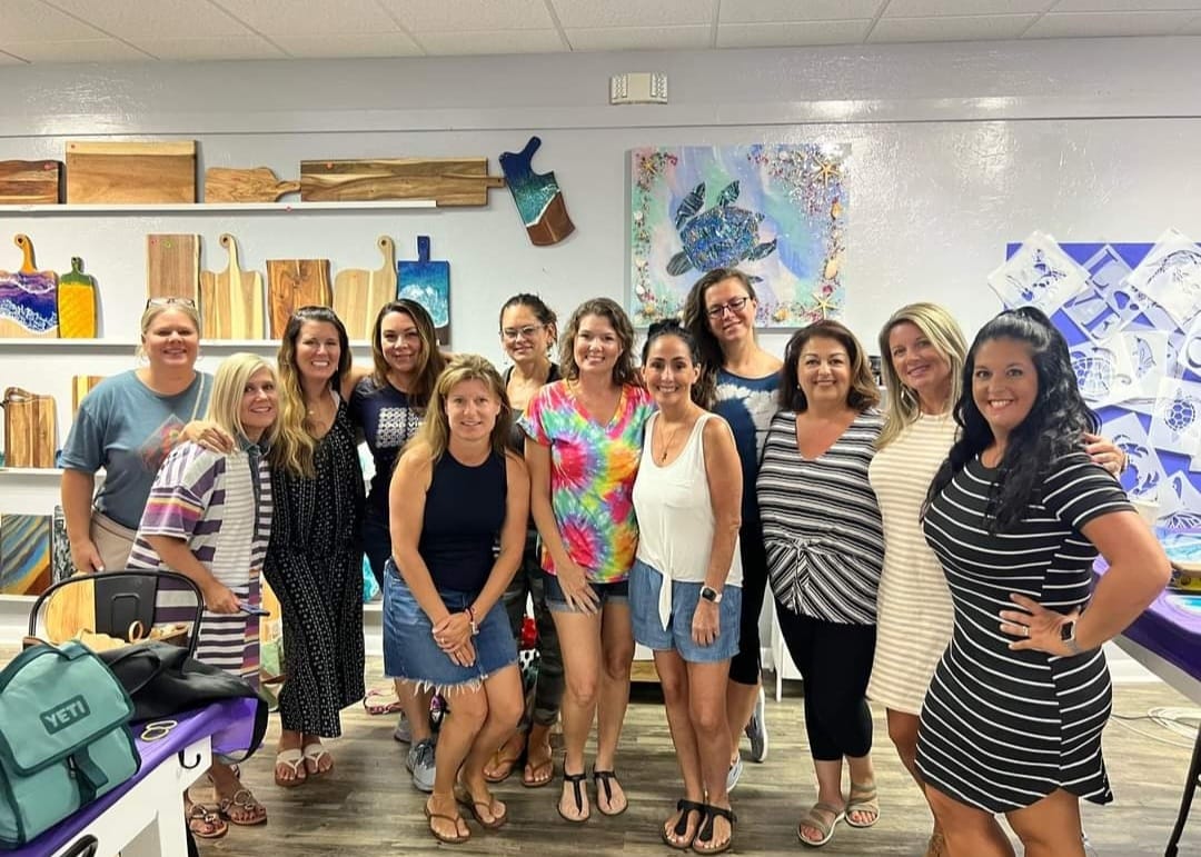 A dozen women smiling and posing in a bright art studio, standing before shelves of wooden cutting boards and colorful ocean-themed wall art featuring a large sea turtle.