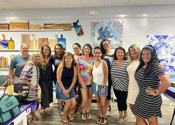 A dozen women smiling and posing in a bright art studio, standing before shelves of wooden cutting boards and colorful ocean-themed wall art featuring a large sea turtle.