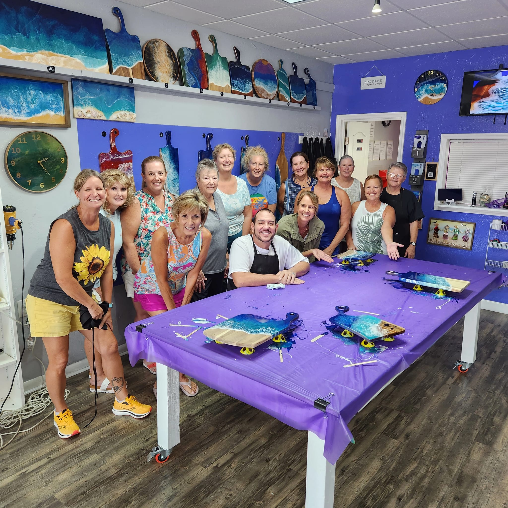 Smiling group of adults in a bright coastal art studio workshop posing around a purple-covered table with ocean-resin serving boards and paddle-shaped wall art