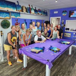 Smiling group of adults in a bright coastal art studio workshop posing around a purple-covered table with ocean-resin serving boards and paddle-shaped wall art
