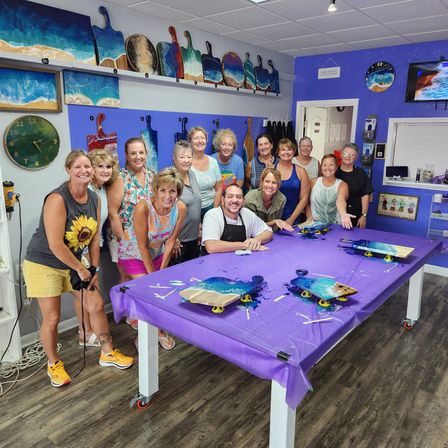 Smiling group of adults in a bright coastal art studio workshop posing around a purple-covered table with ocean-resin serving boards and paddle-shaped wall art