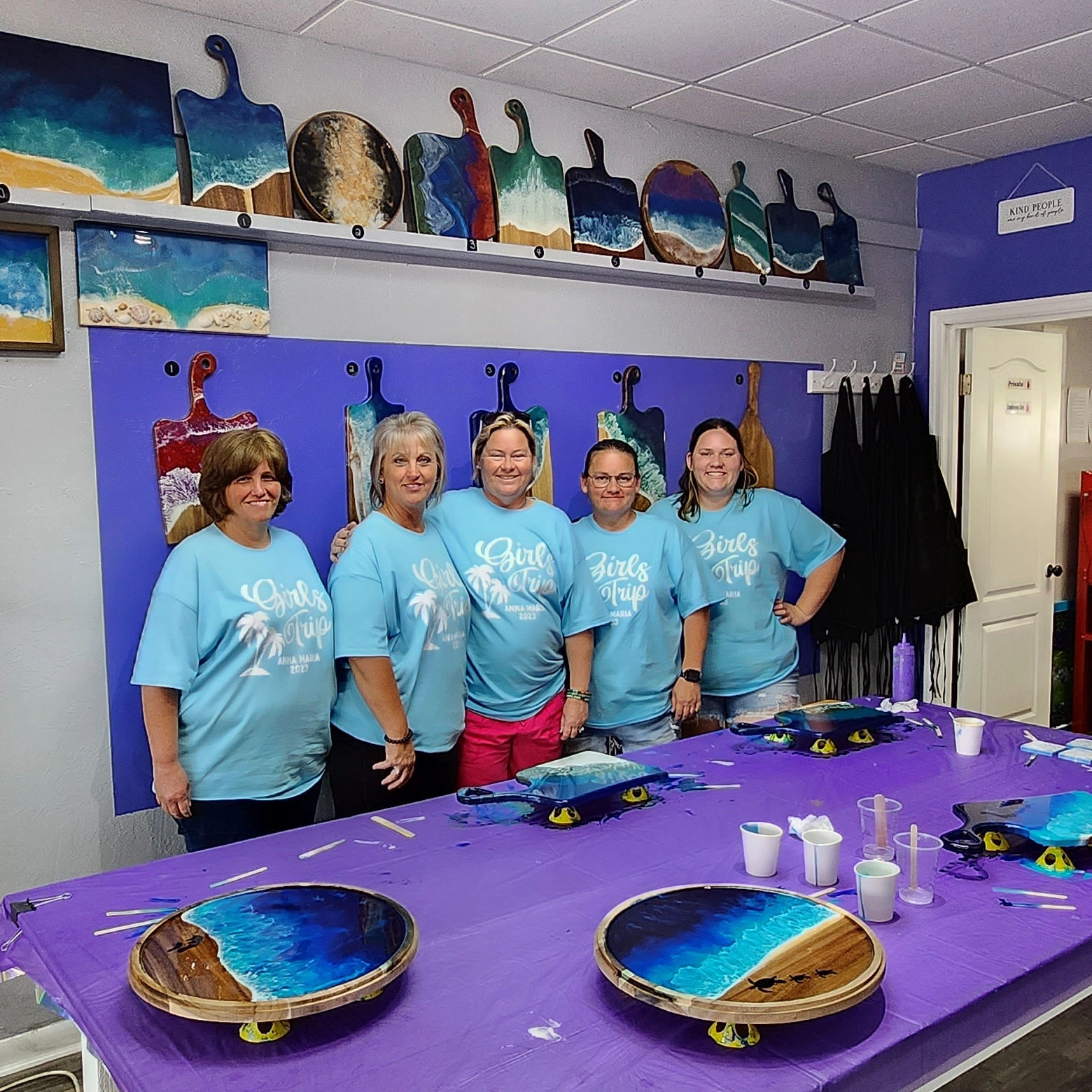 Five women in matching 'Girls Trip' tees pose at an art studio workshop behind a purple table with resin ocean charcuterie boards and paddle-shaped wall art.
