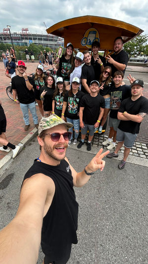 Cheerful group selfie on a yellow party bike at a waterfront promenade with a large stadium and cloudy sky in the background.