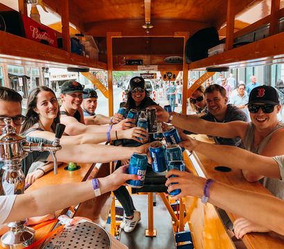 Smiling group clinking beer cans around a wooden bar inside a pedal pub party bike rolling through a busy downtown street, lively bar‑crawl vibe.