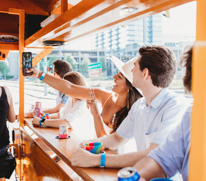 Group of friends riding an orange party bike in downtown, woman in a white hat taking a selfie while others hold colorful cups and drinks.