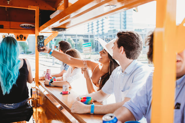 Group of friends riding an orange party bike in downtown, woman in a white hat taking a selfie while others hold colorful cups and drinks.