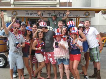 Group of friends wearing red, white and blue outfits and festive Uncle Sam hats, cheering and holding drinks on a Nashville pedal tavern during a patriotic street celebration.