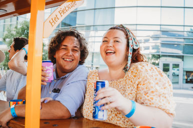 Two friends laughing and holding drinks on a colorful outdoor pedal bar in front of a modern glass building, enjoying a sunny urban outing.