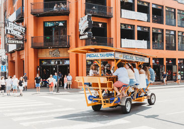 Bright orange pedal-powered party bar with friends pedaling across a downtown Nashville crosswalk past brick music venues and watching pedestrians