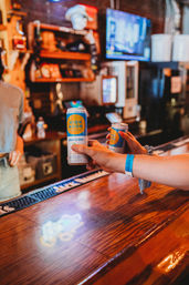 Two hands toasting light-blue and yellow canned vodka & soda over a glossy wooden bar counter in a neon-lit casual bar interior