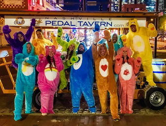 Group of adults in colorful plush bear onesies cheering on a nighttime Nashville pedal bar tour, posing in front of a lit party vehicle on a downtown street.