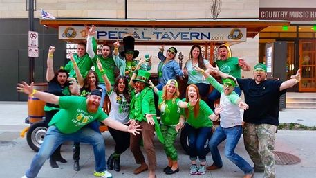 Cheerful group in green outfits and a leprechaun costume posing on a pedal-powered party bike on a downtown street, celebrating St. Patrick’s Day.