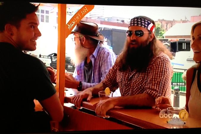 Group chatting at an outdoor pedal-pub style bar on a sunny city street — bearded man wearing an American-flag bandana and sunglasses, an older man with a white beard and hat, and others holding lemon-garnished cocktails.