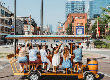Friends riding a bright orange pedal-powered party bar, raising drinks as they roll down a sunny downtown street lined with brick storefronts and glass high-rises.