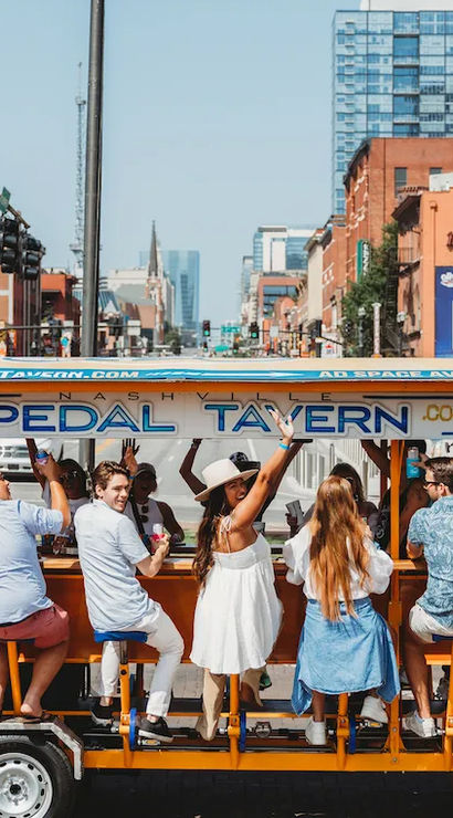 Smiling group pedaling a bright orange party bike through downtown Nashville, passing brick storefronts and modern high-rises on a sunny day.