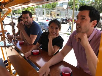 Three people seated at a wooden mobile pedal-bar with red plastic cups, smiling on an urban street with cars and a construction site in the background.