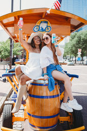 Two friends in summer outfits and sunglasses perched on a wooden barrel of an orange pedal-bar cart on a sunny city street, smiling and raising canned drinks.