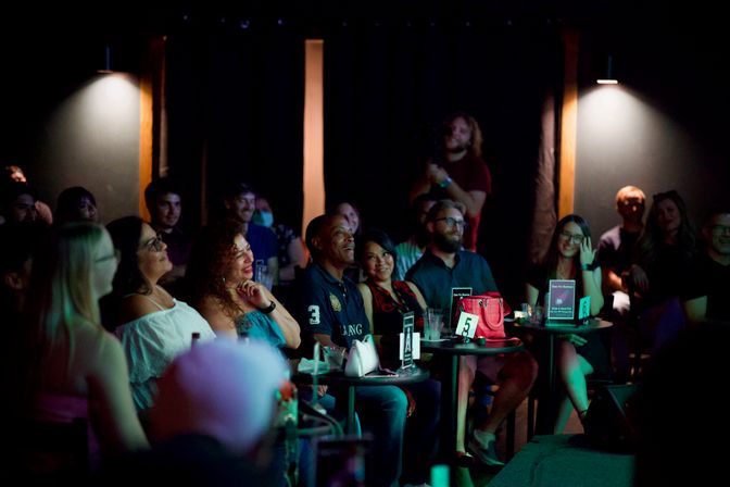 Audience laughing and applauding at a live comedy show in a dim, intimate club — people seated at small round tables with drinks, numbered table cards and stage lighting.