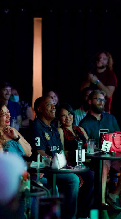 Audience laughing and applauding at a live comedy show in a dim, intimate club — people seated at small round tables with drinks, numbered table cards and stage lighting.