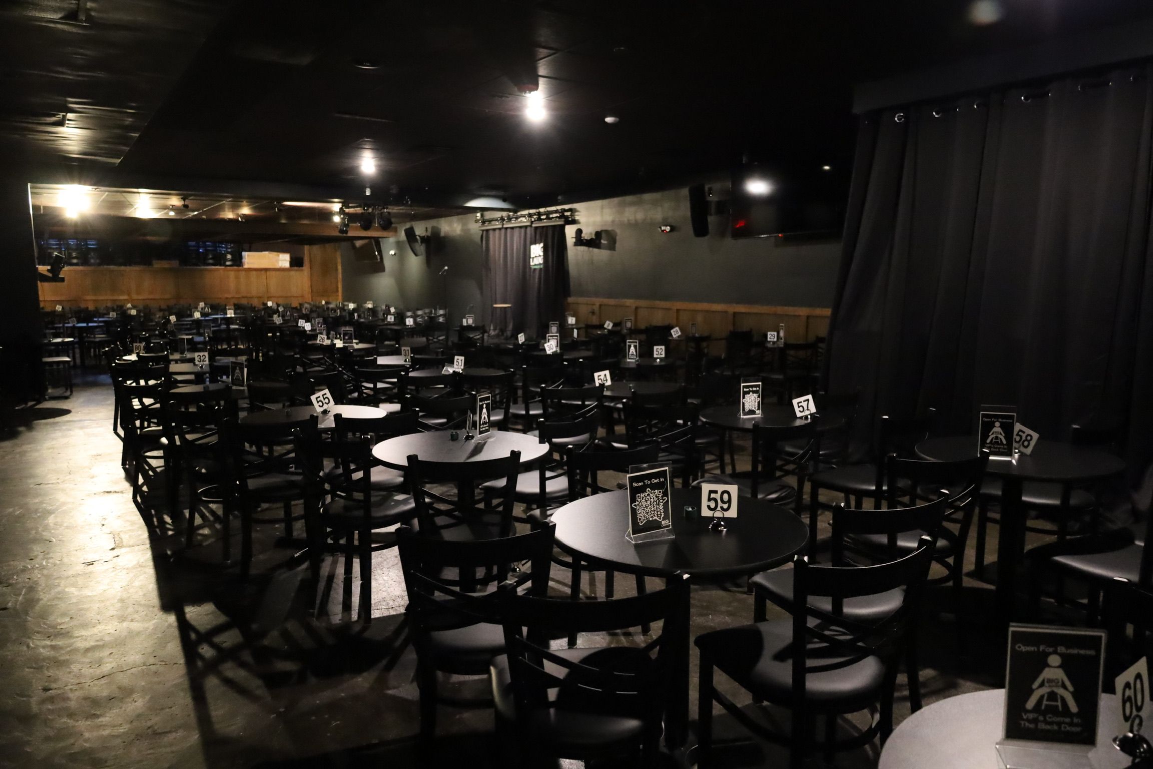 Dimly lit indoor comedy-club style venue with many round tables and black chairs, numbered table signs, and a small curtained stage ready for a show.